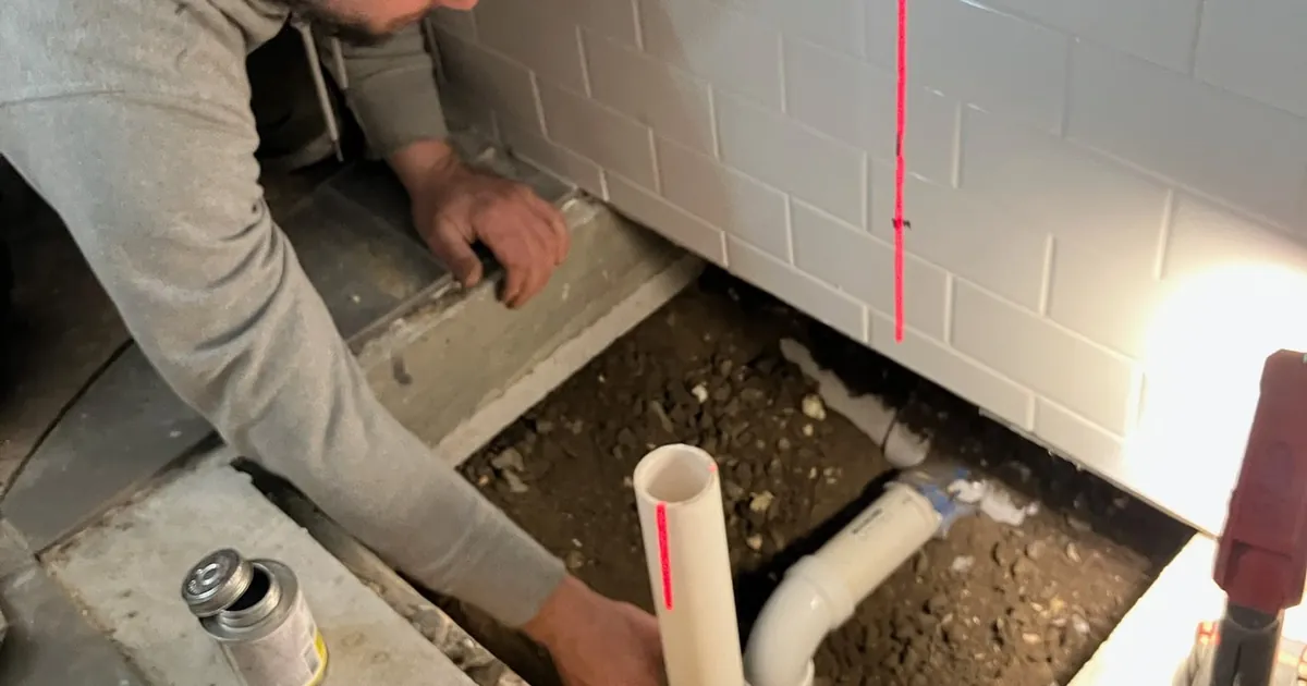 EZ-CO plumber setting a PVC shower drain and trap in a trench cut through a basement concrete slab, aligned with a red laser level